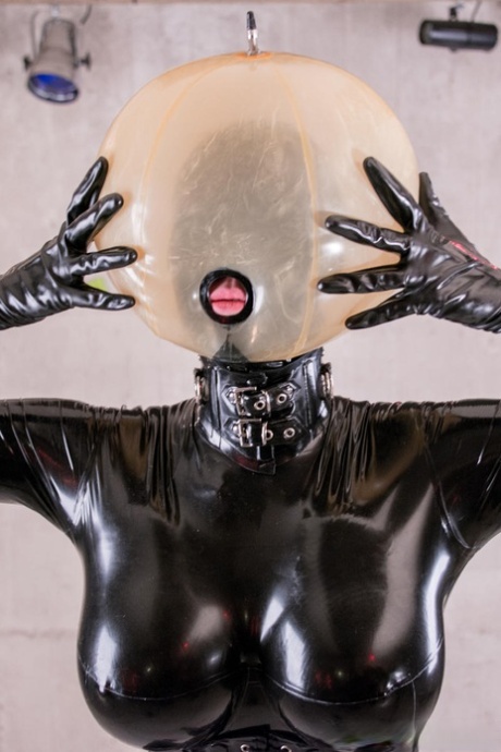 Latex attired slave is affixed with an inflatable hood in dungeon setting
