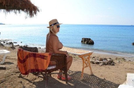 Thick blonde woman with big boobs wanders around a nude beach in a straw hat